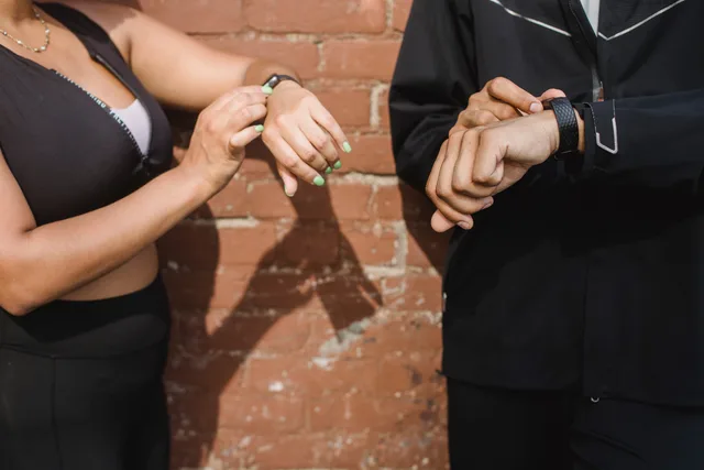 Two people in sportswear checking their fitness trackers outdoors against a brick wall. Fitness creator measurement in England: defining health metrics from the CMO activity guideline